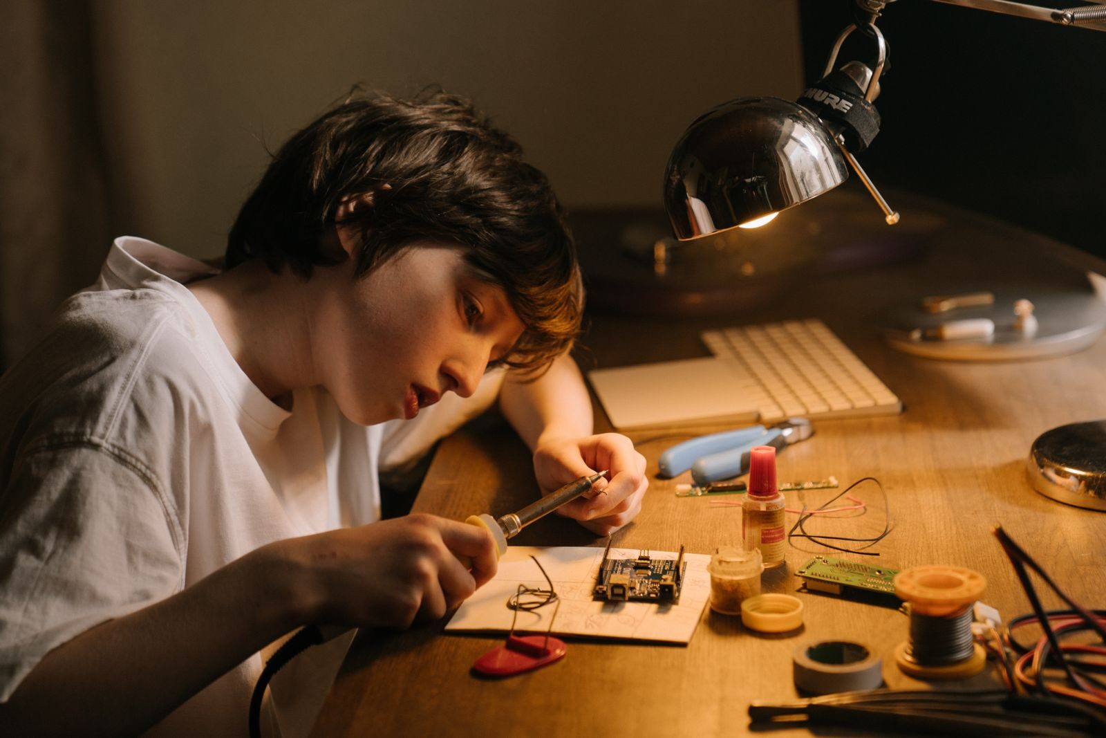Un niño suelda solo una placa de circuito en un escritorio, de noche